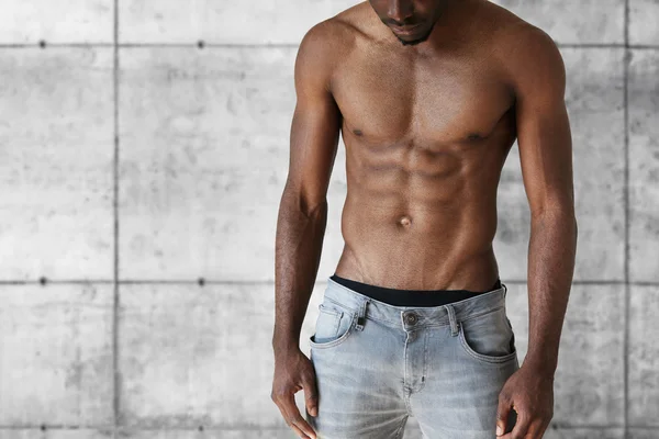 Sport and fitness concept. Cropped shot of handsome well-built athletic young African American man in jeans showing his perfect muscular tense abdominal posing shirtless against brick concrete wall