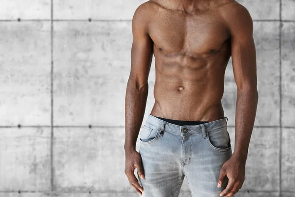 Young dark-skinned model with perfect toned muscular body. Handsome athletic African man in jeans demonstrating his abdominal posing shirtless against gray brick wall. People and lifestyle concept