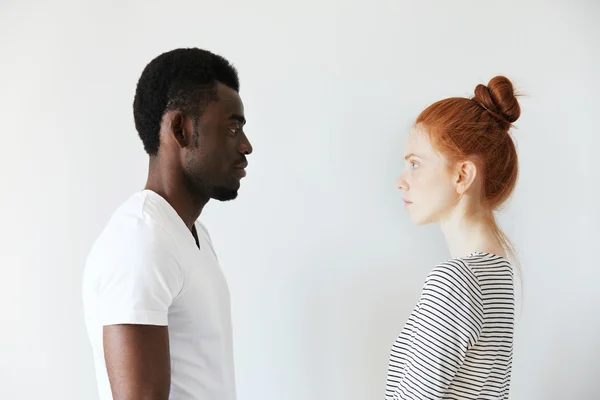 Sideways portrait of a couple looking each other in the eyes on white background. Afro man standing still and Caucasian redhead girl as well watching him with serious look like wife on husband.