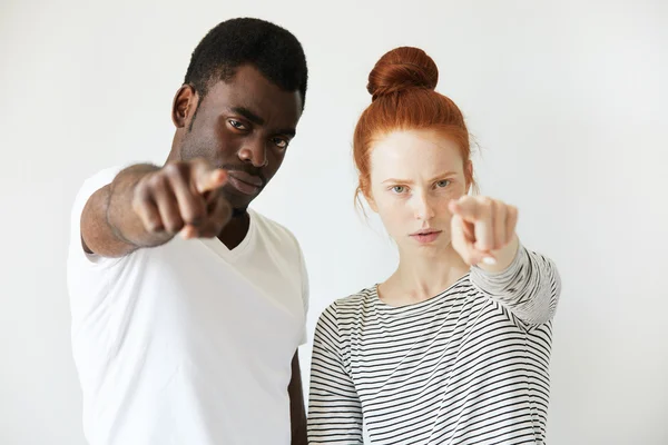 A couple of youngsters pointing index finger in reproach at camera. Mix of Caucasian and African complexions makes this photo looks rich in contrast. Blame and guilty concept. Negative emotions.