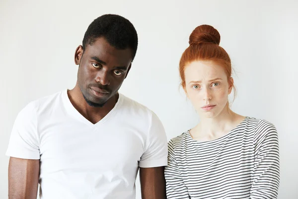Portrait of young Caucasian female with red hair, angry and disappointed with her African boyfriend, who is standing next to her, looking at the camera with tired expression, fed up with quarrelling