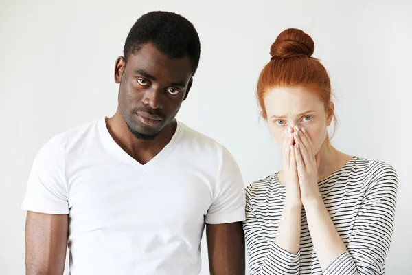 Mixed-race relationships. Interracial couple: Redhead Caucasian woman covering her face and African man in white T-shirt looking at the camera, sorting out their relationships, thinking about divorce.