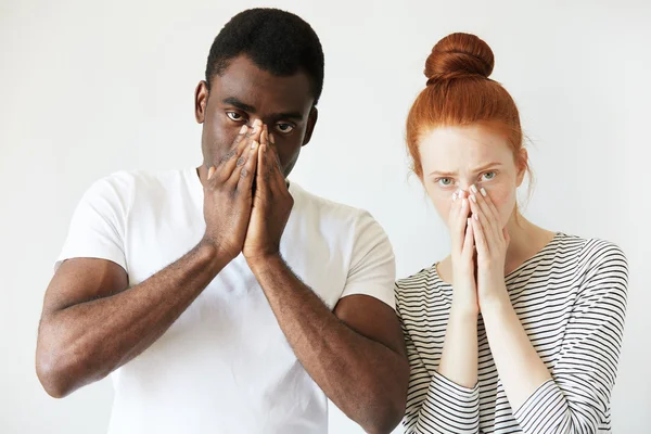 Extraordinary young interracial couple. Caucasian woman in striped top standing next to her African boyfriend looking at the camera with serious expression, covering mouth and nose with both hands