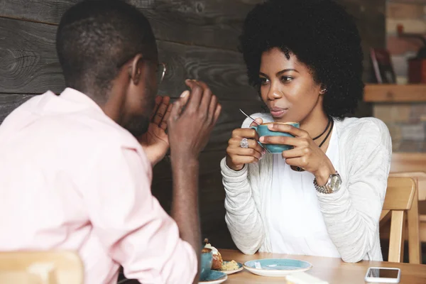 Black girl with Afro hairstyle holding a cup of coffee, listening and looking at her boyfriend with amorous expression. Loving couple enjoying time together at a restaurant on Saint Valentine\'s Day
