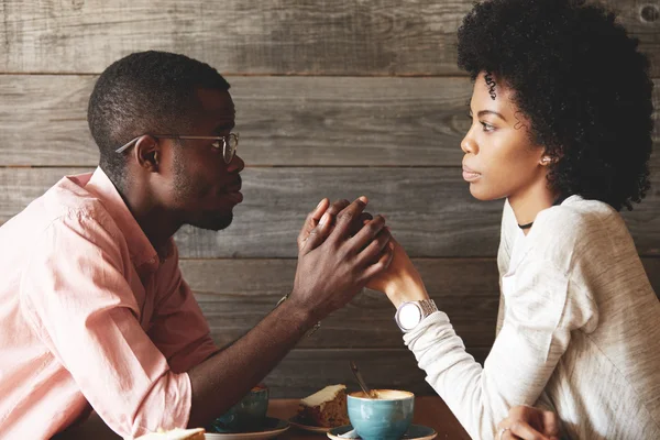 African American young couple sitting at a cafe table: black man in glasses holding her beautiful wife\'s hands, begging her to forgive him, apologizing for an affair, looking with guilty expression