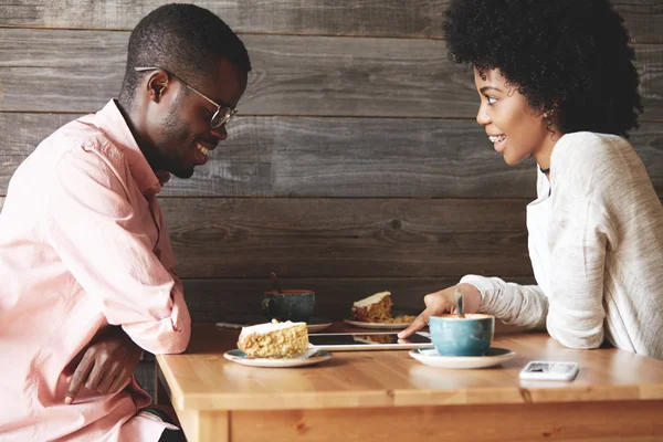 Two African people sitting at a cafe, laughing and having while using tablet together: pretty girl with Afro haircut pointing at the screen, showing something interesting or funny to her friend