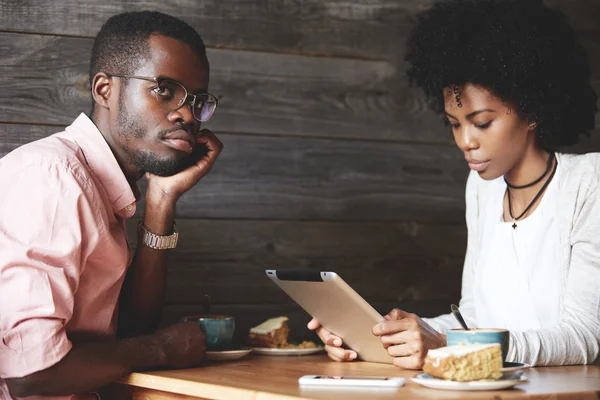 African man with bored and annoyed look, sitting at a cafe with his girlfriend who texting on digital tablet with obsessed expression, using free wireless Internet, totally ignoring her boyfriend