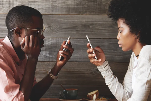 Stylish African couple using cell phones totally absorbed in online life, with obsessed look, not talking to each other, using Wi-Fi at a coffee shop, facing one another. Internet addiction concept