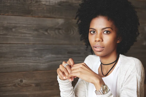 Portrait of fashionable serious young African woman with Afro hairstyle wearing stylish clothes and watches, sitting with crossed hands againts wooden wall background. People and lifestyle concept