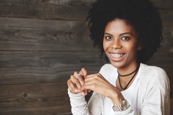 Headshot of stylish dark-skinned female with Afro haircut looking ad smiling at the camera with happy expression, showing braces on upper teeth, against wooden copy space wall for your advertisement