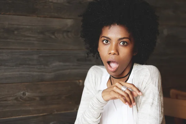 Surprised young black woman, astonished with a gossip or big sale prices, looking at the camera with shocked expression, mouth wide open, against wooden wall with copy space for your advertisement