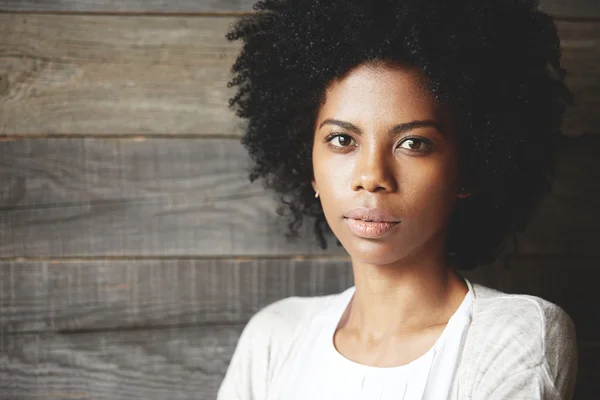 Tender good-looking young African American woman with black curly hair, looking at the camera with brown dreamy eyes, wearing white cardigan, against wooden wall with copy space for your advertisement