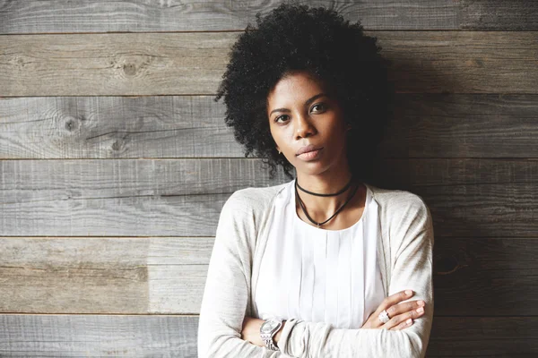 Beautiful African American woman with Afro hairstyle standing with folded arms against wooden copy space wall for your advertising content, looking at the camera with serious expression on her face
