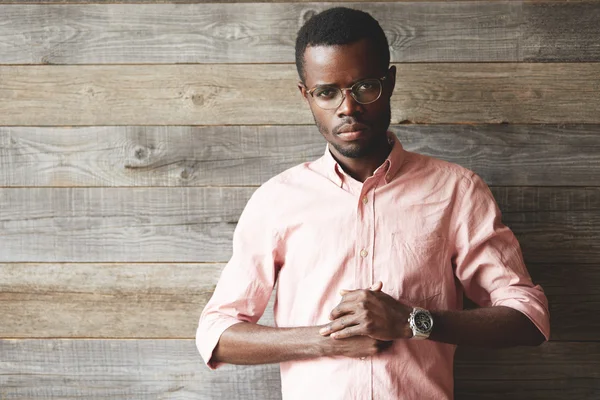 Don\'t play with me. Serious African man in glasses and pink shirt looking at the camera with confident angry expression ready to hit the opponent, holding his hands together against wooden wall