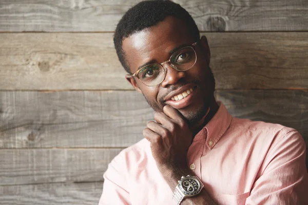 Business and success concept. Close up shot of handsome dark-skinned entrepreneur in glasses and pink shirt, looking at the camera with happy confident smile showing white teeth, holding his chin