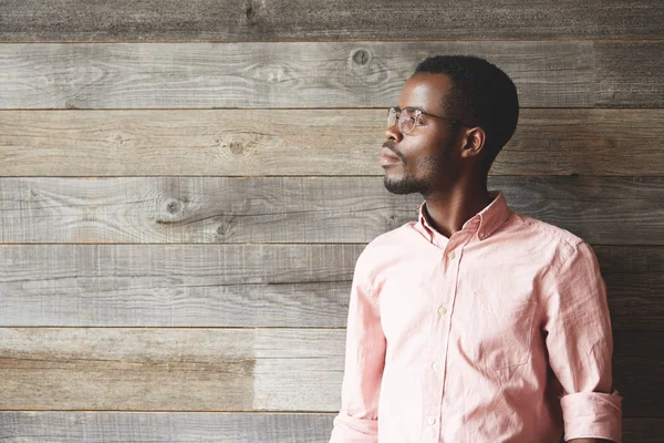 Profile portrait of stylish African American businessman in pink shirt and spectacles, looking away with thoughtful expression against wooden wall with copy space for your text or advertising content