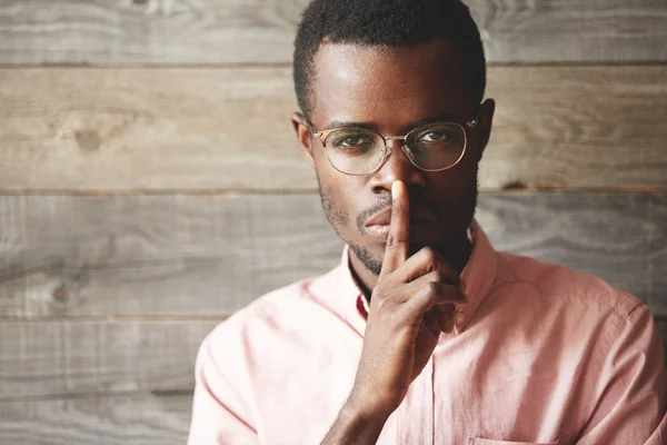 Top secret. Young African businessman in pink shirt holding a finger on his lips, asking to keep silence about confidential information, saying 'shh', looking at the camera with serious expression.