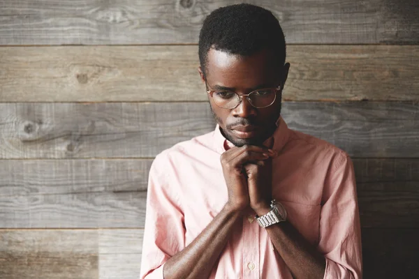 Close up shot of handsome young African man praying and meditating against wooden background. Dark skinned student wearing pink shirt, holding hands in prayer against his lips, hoping for the best