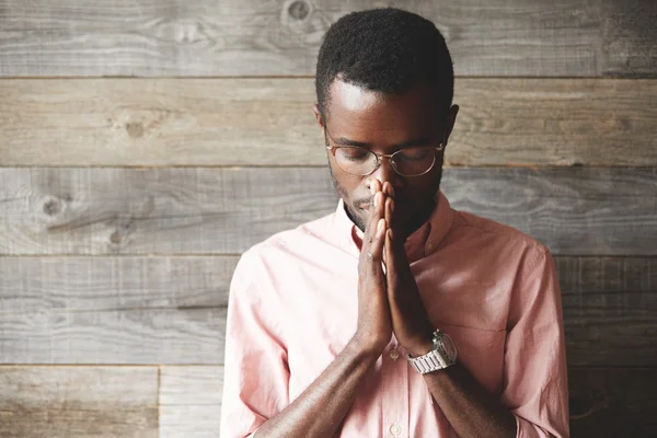 Young meditating and praying African American man wearing pink shirt and glasses, holding hands in prayer against his lips, hoping for the best, asking for wisdom and strenght, with closed eyes