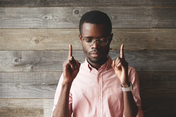 Portrait of handsome young African man, standing against wooden wall background, with closed eyes, showing index fingers up, trying to remember something very important, looking concentrated