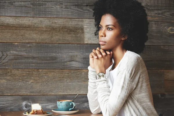 Young attractive dark skinned female at a cafe table, drinking coffee with cake, sitting in thinking pose, resting chin on her clasped hands, looking away with a reflective expression on her face