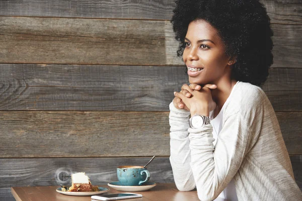 Close-up profile portrait of African girl, having braces on upper teeth, smiling to her boyfriend who is entering the cafeteria, resting chin on her hands. Mug, cake and mobile phone on the table