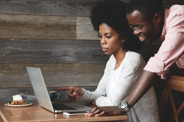 Stylish African female pointing a finger at the screen of generic laptop showing something funny to her boyfriend who is standing next to her and laughing. Team work: two colleagues working at a cowor
