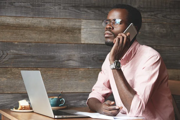 Successful African businessman in glasses and pink shirt talking on the phone to his partner, listening attentively, looking into distance with pensive expression, sitting at a coffee shop during lunc