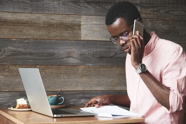 Young African executive officer in formal shirt examining a financial report, making phone calls to his accountant for specifying details and figures, while working on laptop sitting at a coffee shop