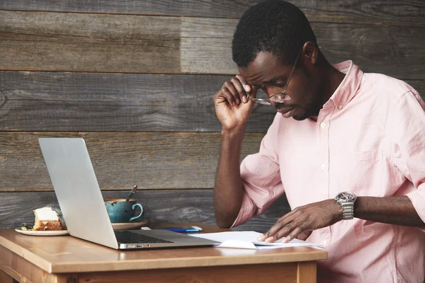 Young black estate agent studying details of a lease contract, looking concentrated, perusing papers through his glasses, sitting at a cafe in front of laptop, mug and pie in pink shirt and watches