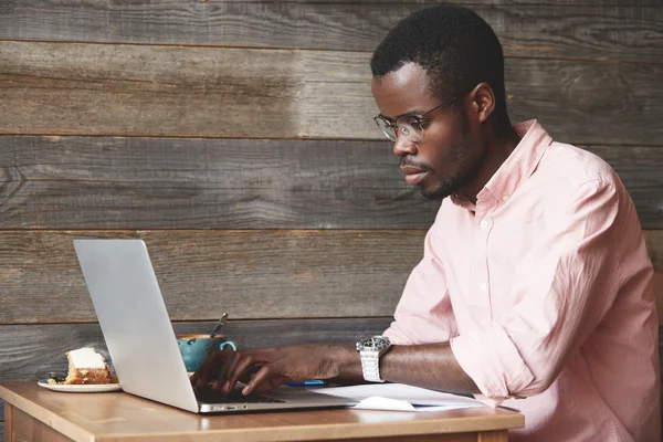 Profile portrait of young African web shop manager, typing a message on a laptop to his partner about changes in delivery terms of a new collection of clothes, isolated on wooden background of a cafe