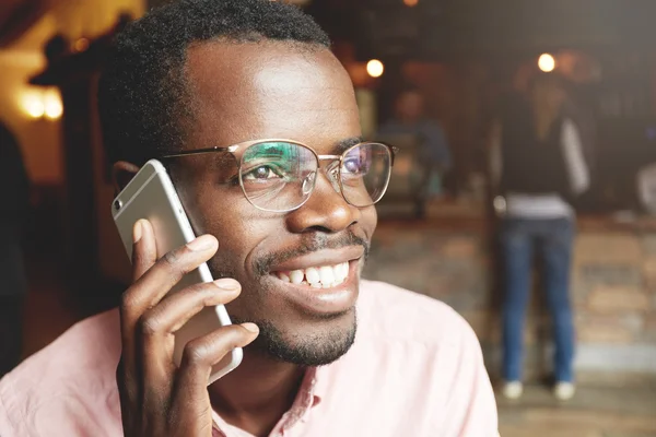 Happy and cheerful young African man in glasses and pink shirt, with joyful smile and amorous glance on his cute unshaven face, talking on mobile phone with his girlfriend, sitting at a coffee shop