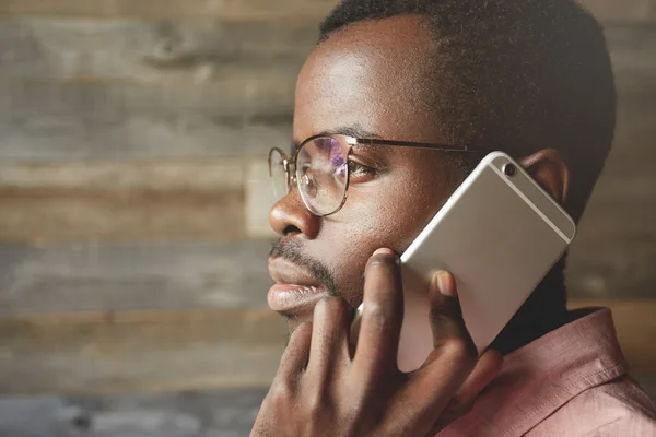 Profile portrait of attractive black man wearing spectacles looking through the window while listening to messages on his mobile phone, with thoughtful and calm face expression against wooden wall