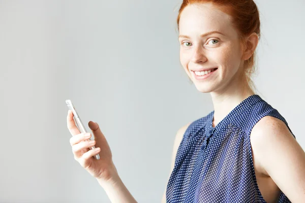 Young red haired Caucasian female posing with new model of smart phone in her hand, looking at the camera with happy smile, against white wall with copy space for your advertising information