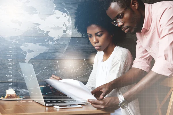 Team work and cooperation. African corporate worker in shirt and glasses holding papers, showing a report to his female colleague with Afro hairstyle, working on laptop computer. Visual effects