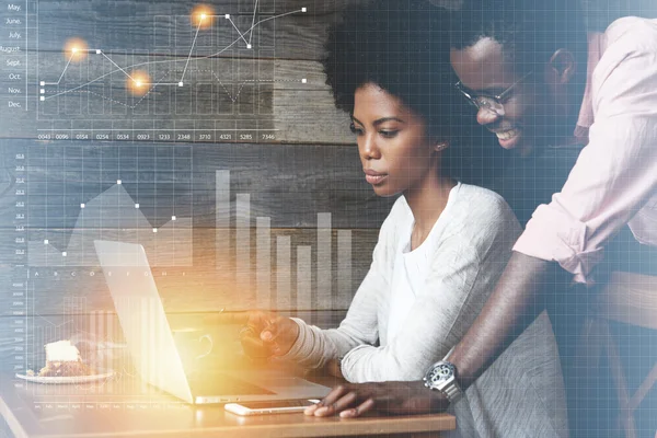 Happy African freelancer watching his female partner with Afro haircut showing graphics, demonstrating growth rates in sales, pointing a finger at the laptop screen. Double exposure. Visual effects