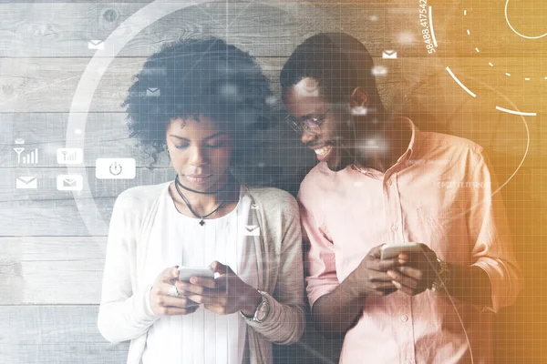 Worldwide connection interface. African couple using gadgets: smiling man holding mobile phone looking over his shoulder at the screen of his girlfriend\'s smart phone, viewing photo on the Internet
