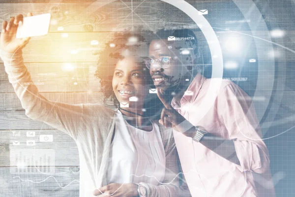 Worldwide connection interface. Young happy African American couple having fun, taking selfie with futuristic cell phone, smiling at the camera, posing against wooden wall background. Visual effects