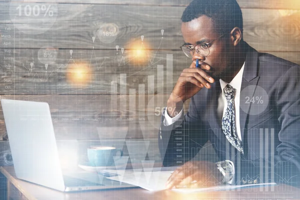 Visual effects. Serious African businessman in spectacles looking at the laptop screen with thoughtful expression, holding a pen, thinking over his business ideas, doing paperwork, having cappuccino