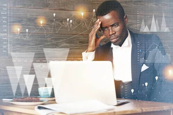 Visual effects. Double exposure of tired African businessman in glasses using laptop computer with concentrated look, touching his head, having bad headache while working late hours in the office