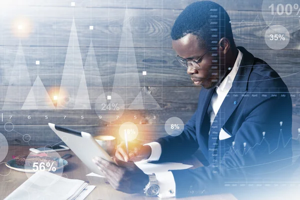 Double exposure of African businessman in elegant suit and glasses filling in a financial report holding a pen in one hand and digital tablet in the other. Graphics, diagrams interface. Film effect