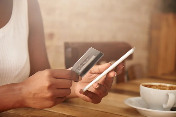 Selective focus. Close up shot of female hands holding plastic card and cell phone. African woman paying with credit card while shopping online, using mobile phone, having cappuccino at a restaurant