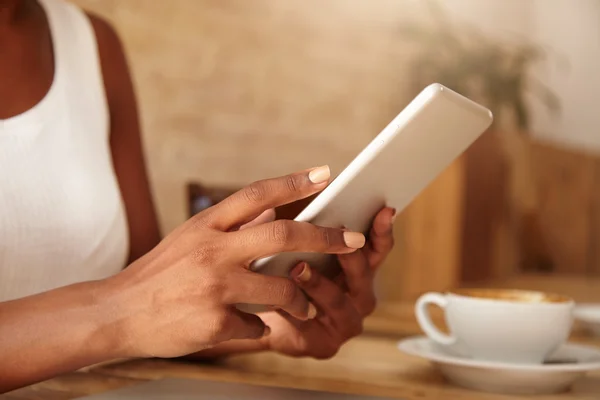 Croped portrait of young dark-skinned girl in white tank top, sitting at the cafe table, typing on her white tablet with her thin manicured fingers, while drinking coffee. Selective focus on hands