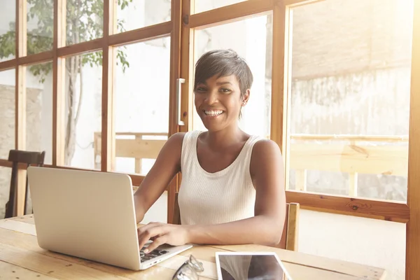 Young smiling dark-skinned female with short haircut sitting at the wooden table in front of notebook, browsing online-shops, dressed casually, looking at the camera with happy face expression