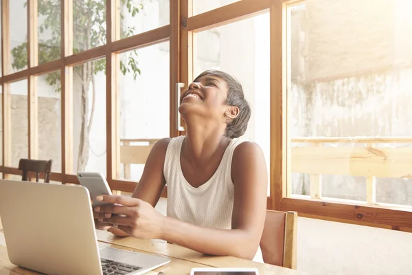 African student girl laughing out loud using smartphone, chatting with her friends online, having fun, sitting at a cafe with notebook with blank copy space for your advertising content on the table