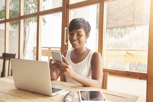 Young black woman answering a call on Internet-enabled smart phone while working on tablet and notebook with blank copy space for your promotional information sitting against large windows background