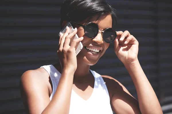 Pretty African office worker chilling in the shadow wearing sunglasses during coffee break. Black female talking on mobile phone, smiling with happy and carefree look, touching her stylish shades