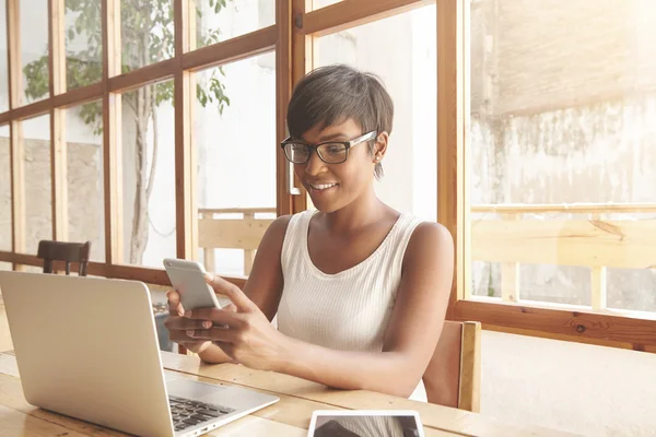 Indoor portrait of black businesswoman in glasses using wireless Internet connection on laptop and gadgets, messaging via popular social networks, dressed casually. People and technology concept
