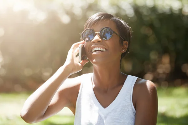 Half profile of beautiful young woman wearing casual white top and stylish shades laughing while talking on cell phone with happy look and mouth wide open, having a picnic in the park on sunny day