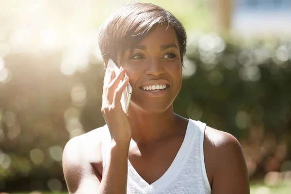 Close up shot of African American student girl with short hairstyle talking on mobile phone with happy excited look while having a picnic with her friends in the public garden on sunny summer morning